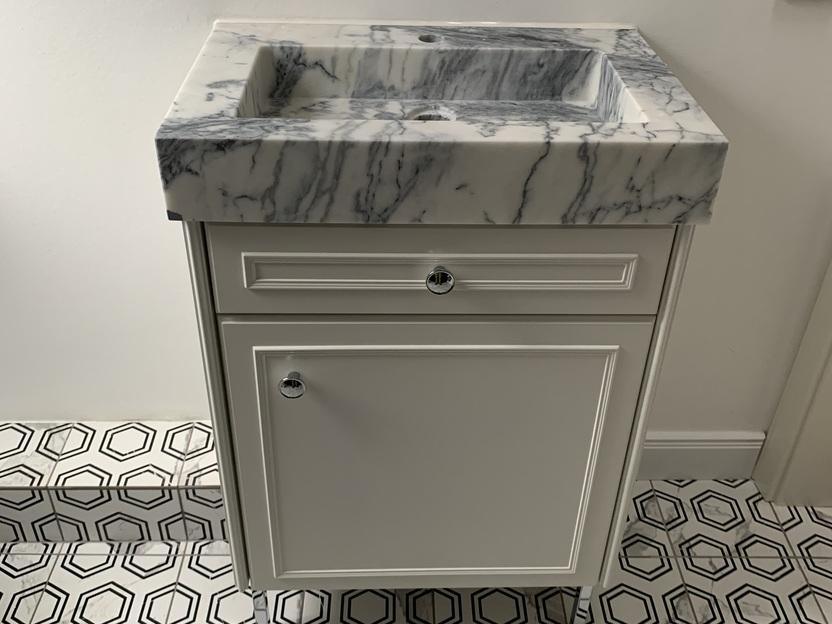 Maßgefertigter Waschtisch mit Marmor-Waschbecken Ein kompakter, maßgefertigter Waschtisch mit einem edlen Marmor-Waschbecken auf einem weißen Unterschrank. Der Boden ist mit schwarz-weißen, geometrischen Fliesen gestaltet. Badmöbel nach Maß.