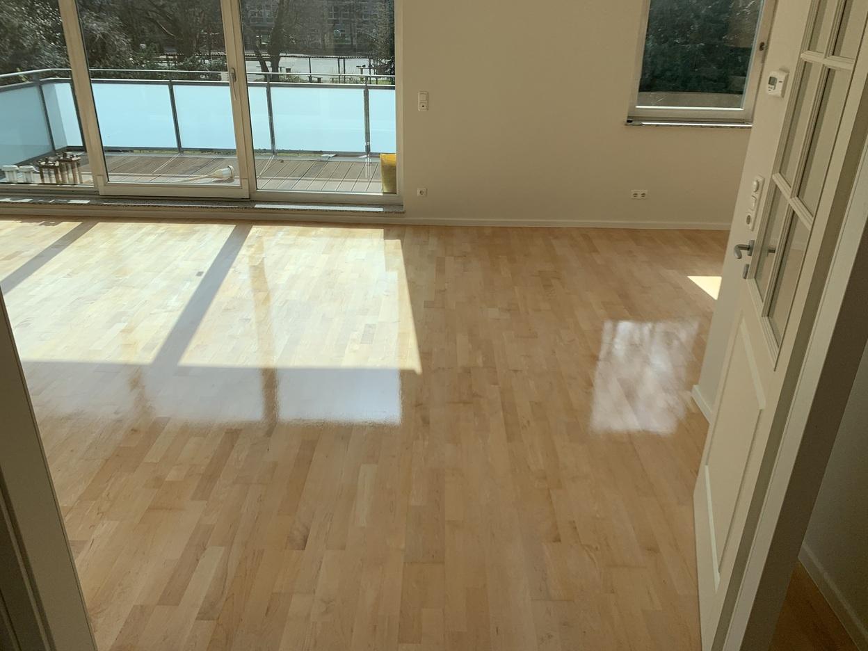 Ein heller, glänzender Holzboden in einem lichtdurchfluteten Wohnraum mit Zugang zu einem Balkon. Der Boden wurde individuell gestaltet und verlegt. Individueller Boden.