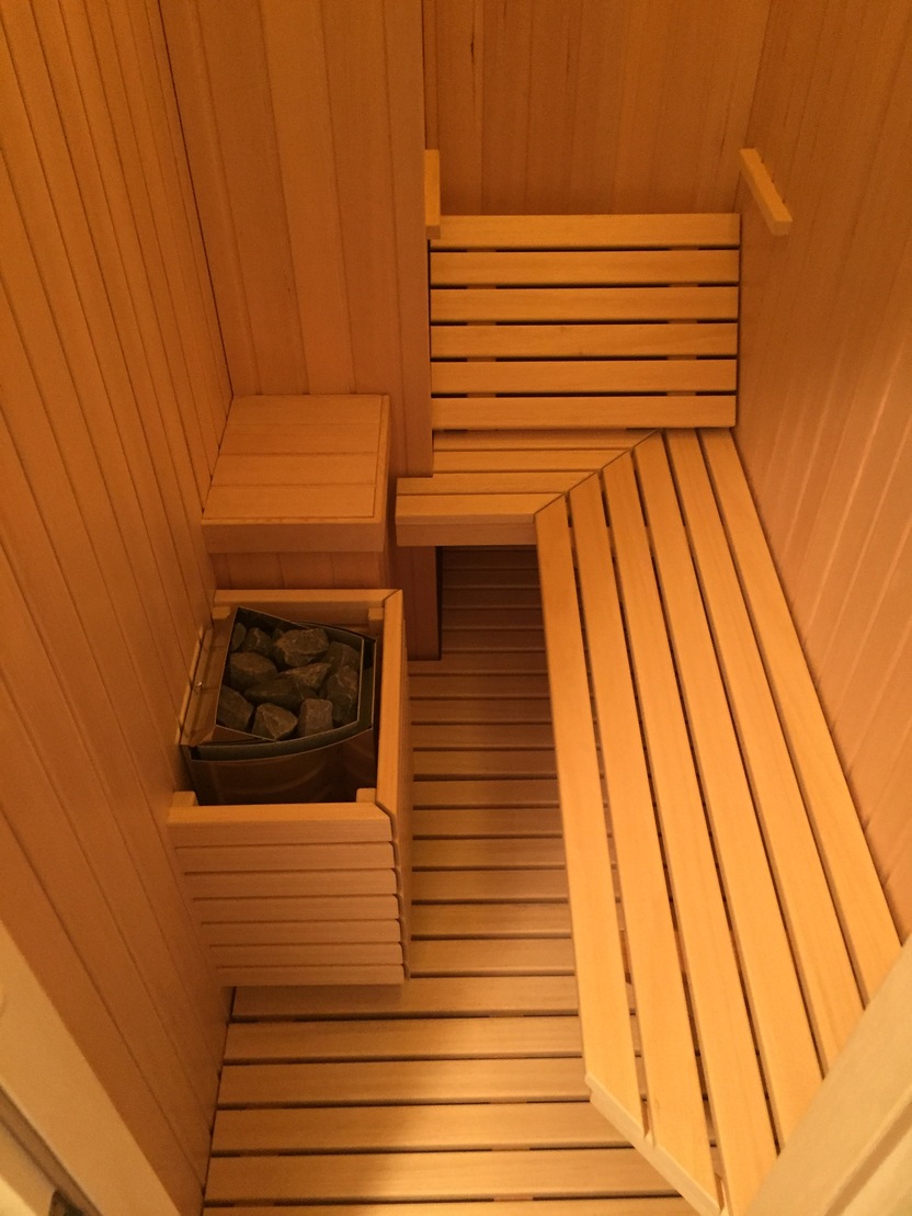 Blick von oben in eine kleine Sauna mit L-förmiger Sitzbank, holzverkleideten Wänden und Bodenrost, seitlich platzierter Saunaofen mit Schutzgitter