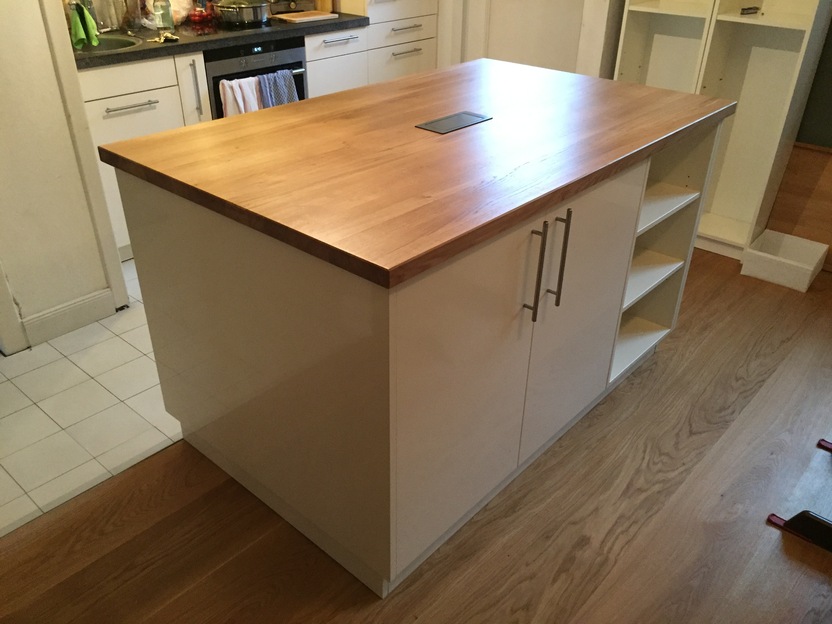 Kücheninsel mit heller Front, integrierten Regalfächern und einer massiven Arbeitsplatte aus Holz, im Hintergrund klassische Einbauküche