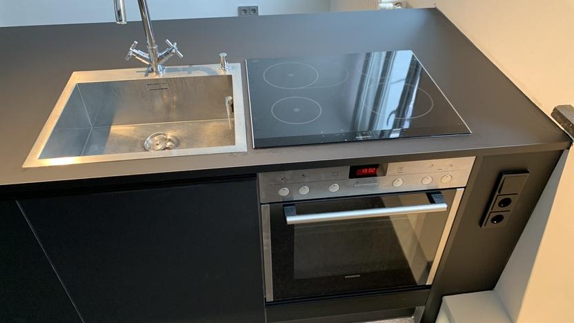 Bild einer modernen Kücheneinheit mit einer schwarzen Arbeitsfläche. Die Einheit beinhaltet ein Edelstahlspülbecken mit einem hochglanzpolierten Wasserhahn und ein integriertes schwarzes Induktionskochfeld.