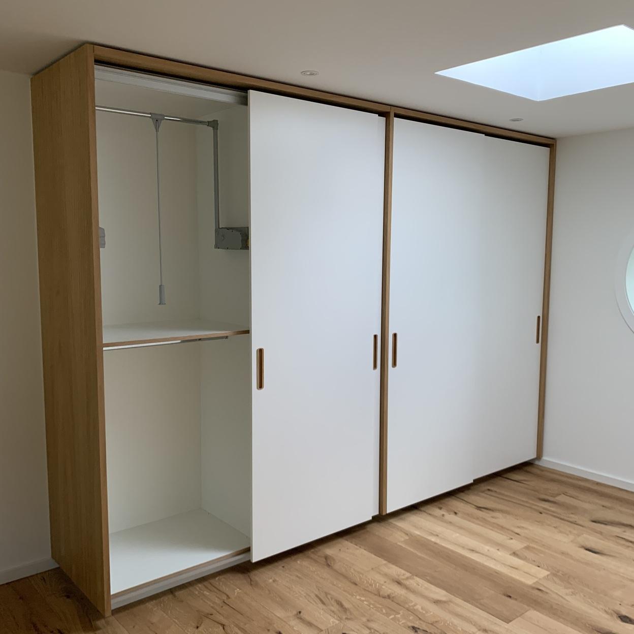 Schiebetürschrank mit offenem Fach und Kleiderlift Maßgefertigter Schrank mit drei weißen Schiebetüren und geöffneter Abteilung links, darin ein ausziehbarer Kleiderlift und eine Kleiderstange, kombiniert mit hellem Holzdekor und Griffmulden