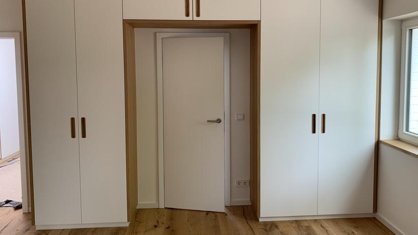 Maßgefertigter Einbauschrank um eine Tür Ein maßgefertigter Einbauschrank in einem modernen Design, der eine Tür umrahmt. Der Schrank hat weiße Fronten mit Holzgriffen und passt perfekt in den Raum. Möbel nach Maß.
