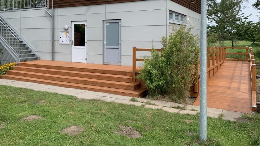 Maßgefertigte Terrasse mit Stufen und Geländer vor einem Jugendzentrum Eine maßgefertigte Terrasse mit breiten Stufen und Holzgeländer vor einem Jugendzentrum. Ein farbenfrohes Banner hängt an der Wand des Gebäudes. Terrasse nach Maß.