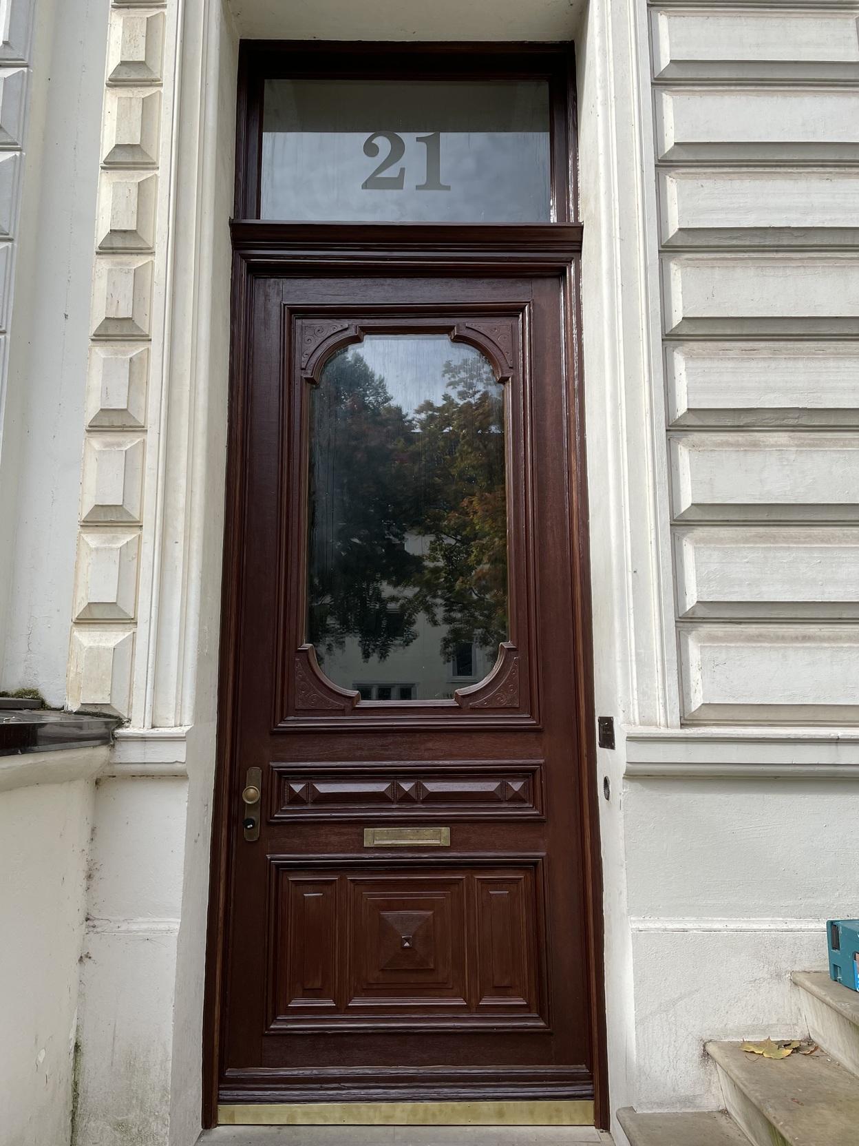 Eine dunkelbraune, ma&szlig;gefertigte Haust&uuml;r aus Holz mit dekorativen Elementen und einem gro&szlig;en Glaseinsatz. &Uuml;ber der T&uuml;r befindet sich die Hausnummer 21. Haust&uuml;r nach Ma&szlig;.