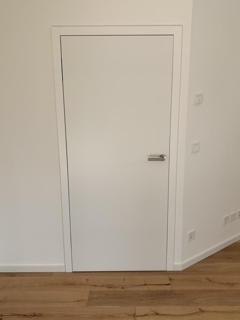 Maßgefertigte Zimmertür in modernem Design Eine schlichte, weiße maßgefertigte Zimmertür mit Edelstahlgriff fügt sich nahtlos in die Wand ein. Der Boden besteht aus Holz und betont das minimalistische Design. Zimmertür nach Maß.